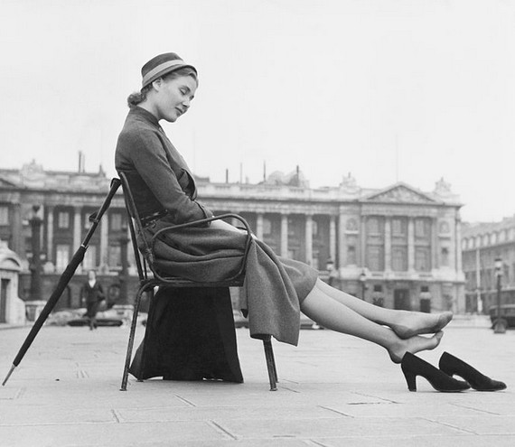 Woman in dress and hat sitting on a chair by a square and has taken off her shoes. An umbrella is leaning against the back of the chair, 1950s Woman in dress and hat sitting on a chair by a square and has taken off her shoes. An umbrella is leaning against the back of the chair, 1950s